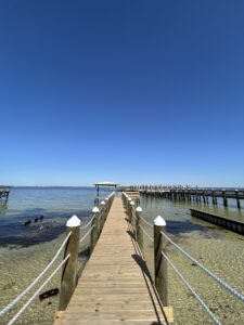 Wooden dock at The Alice stretching over clear emerald bay waters under a bright blue sky.