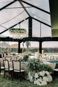 Interior of a clear-top tent at The Alice set with long banquet tables, floral arrangements, and a hanging chandelier overlooking the lawn and palm trees.