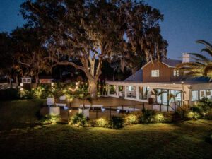 Evening view of The Alice’s illuminated deck and venue building under a large oak tree with string lights and landscaped gardens.